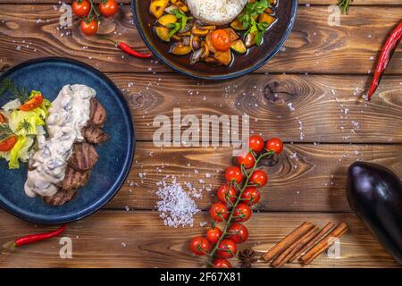 Beef patty on a platter with vegetables, stewed vegetables with rice, lettuce, bell pepper, rosemary, garlic and sesame on a brown wooden background. Stock Photo