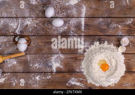 Chicken eggs, a pile of wheat flour and a rolling pin on a brown board background. The process of preparing the dough for baking. Flat layout. Stock Photo