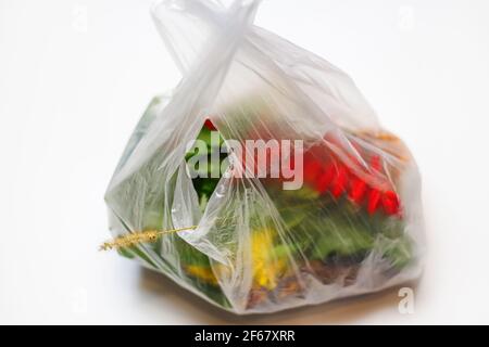 DEFOCUS. Plastic bag. Red and green plants flowers in a plastic bag on a white background. A dry blade of grass sticks out. Ecological problems. Out o Stock Photo