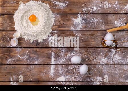 Chicken eggs, a pile of wheat flour and a rolling pin on a brown board background. The process of preparing the dough for baking. Flat layout. Stock Photo