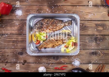 Grilled fish with vegetables, bell pepper, coarse salt, laid in an aluminum container on a brown wooden background. Restaurant food delivery. Stock Photo