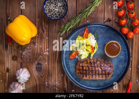 Beef patty on a platter with vegetables, lettuce, bell pepper, rosemary, garlic and sesame seeds on a brown wooden background. Restaurant service. Stock Photo