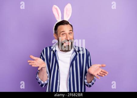 Photo of optimistic mature man in theme easter rabbit ears headband shrug shoulders isolated over violet color background Stock Photo