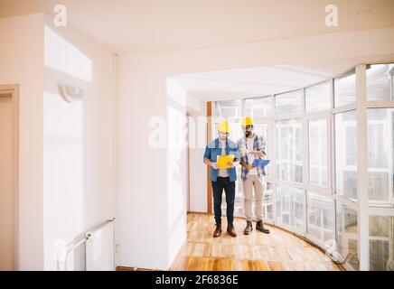 Two focused innovative young engineers men with helmets and folders sharing ideas in the bright office. Stock Photo