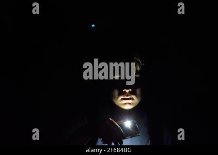 Young Man in a Dark Cave Shining His Phone's Torch Under His Face Stock Photo