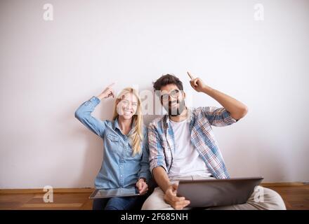 Happy charming young love couple pointing on the empty space above while sitting on the floor. Stock Photo