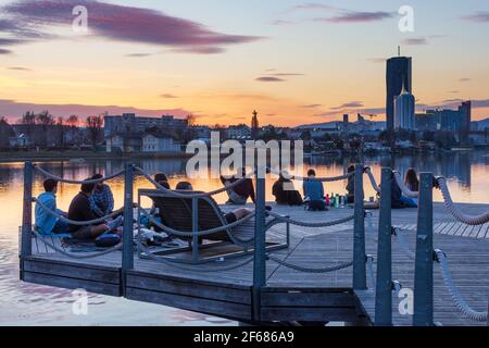 Wien, Vienna: young people on a platform at a party above oxbow lake Alte Donau (Old Danube) at fiery sunset, buildings of Donaucity, DC Tower 1, Coro Stock Photo