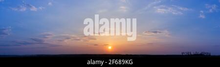 Panorama of blue and orange sunset, sky with clouds, evening over the field. Silhouettes of trees in the valley. Stock Photo