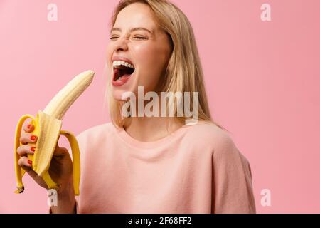 Joyful charming blonde girl laughing and eating banana isolated over pink background Stock Photo