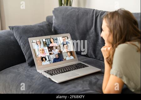 Laptop screen with video call on it. Young woman using computer app for video communication with diverse group of friends or colleagues, female employee works remotely from home, lying down on couch Stock Photo