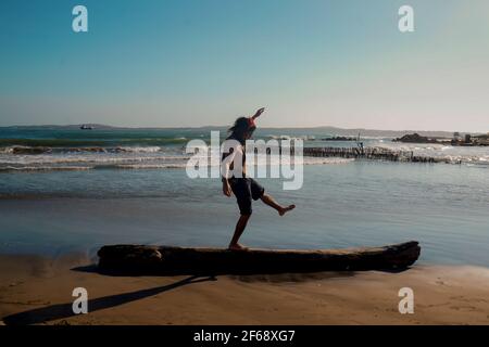 Man with long hair walking and playing on a piece of wood on the beach Stock Photo