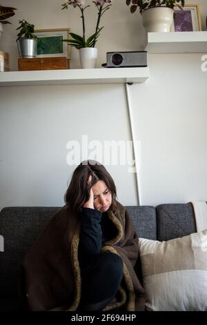 Woman sitting on couch under blanket with head in hand Stock Photo