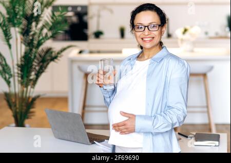 Healthy pregnant female. Joyful pregnant mixed race woman, leads a healthy lifestyle, holds a glass of clean water, drinks the daily rate of water, looks at the camera, smiles, care about future baby Stock Photo