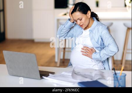 Upset stressed pregnant mixed race adult woman, designer, manager or freelance works from home, dressed in stylish wear. Pregnant lady suffering from headache, overworked, touches head and belly Stock Photo