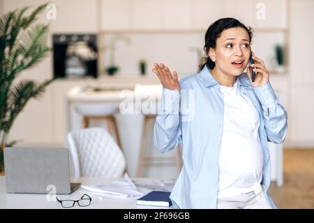 Outraged pregnant busy woman. Stressed irritated mixed race pregnant woman, business lady or office coworker, stylishly dressed, standing near the table, indignantly talking on the phone Stock Photo