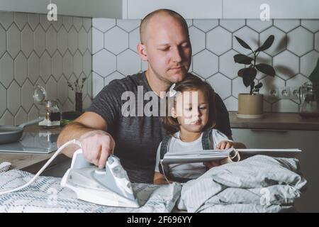 Man ironing bed linen and reading book to his little daughter. Father engaged in household chores. Stock Photo