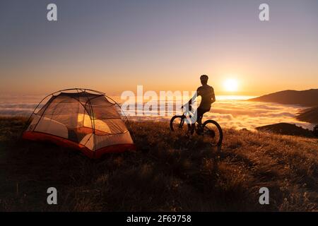 Mountain biker standing next to tent at sunset enjoying the view Stock Photo