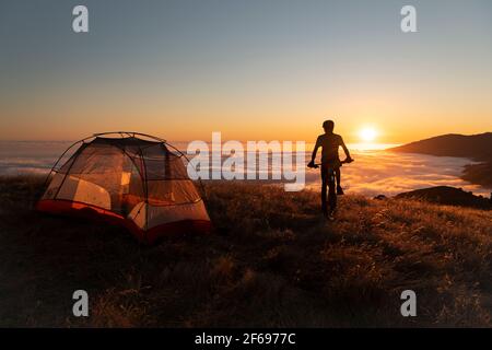 Mountain biker standing next to tent at sunset enjoying the view Stock Photo