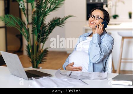Elegant smart confident pregnant brunette mixed race female business woman, designer or freelancer, working at home using laptop, talking on the cellphone with colleague or client, smiling Stock Photo