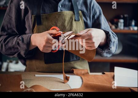 A young tanner in his workshop at the table cuts out leather elements of shoes. Stock Photo