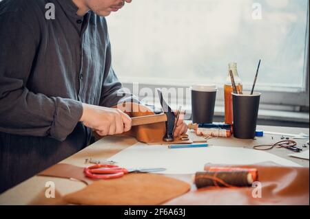 A young shoemaker using a hammer knocks down rivets on a leather product made with his own hands. Stock Photo