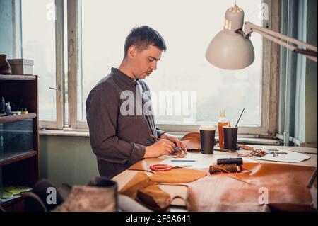 A young apprentice helps his father, glues small leather parts for shoes with a brush in the family workshop. Stock Photo