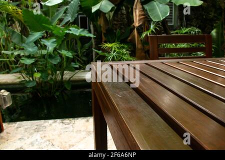 The corner of a mahogany wood slat table and a matching chair on a veranda surrounded by a moat and thick jungle foliage in the Caribbean. Stock Photo