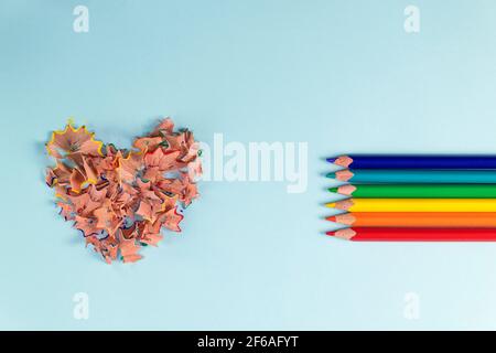 sharpened colored pencils and heart-shaped pencil shavings on pastel blue color. Rainbow or LGBT pencils. Decoration for St. Valentine's Day. Top view