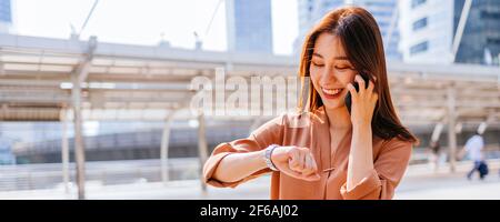 Successful young Asian woman in formal clothing checking time in wristwatch while walking and talking over phone in city street at night Stock Photo