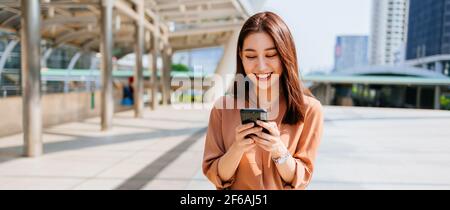 Beautiful young Asian woman in formal clothing standing outside office building using smartphone while browsing internet during break Stock Photo
