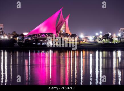 Panoramic view of the distinctively sail-shaped illuminated clubhouse of Dubai Creek Golf and Yacht Club from across the Dubai Creek park. Stock Photo