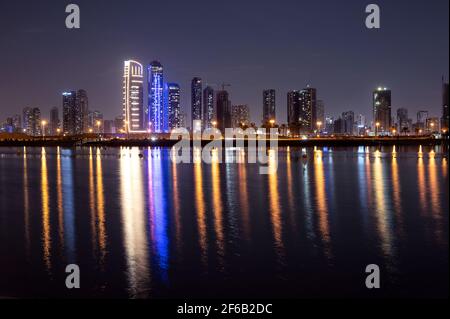 Sharjah night skyline with illuminated sky scrappers,buildings showing reflection on water along with moving clouds captured from Mamzar Beach Stock Photo