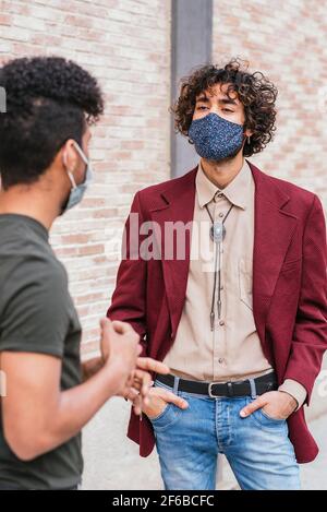 portrait of a caucasian young man dressing trendy clothes and blue face mask. He is talking with latin colleague in the street near a brick wall Stock Photo