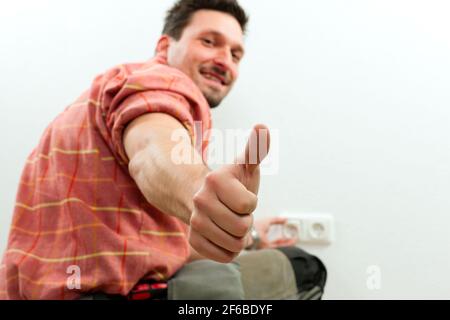 Electrician installing a power socket giving the thumbs-up sign - focus on thumb Stock Photo