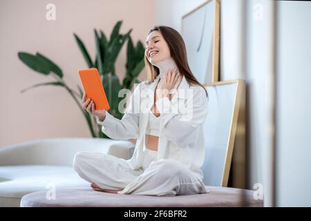 Pretty woman in white sitting on a soft chair and making a photo of tapes on her neck Stock Photo