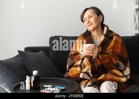 Smiling senior woman feels relieved, recovering after viral disease. An elderly female covered blanket sits on the couch, drinks hot and feels better. Healthcare concept Stock Photo