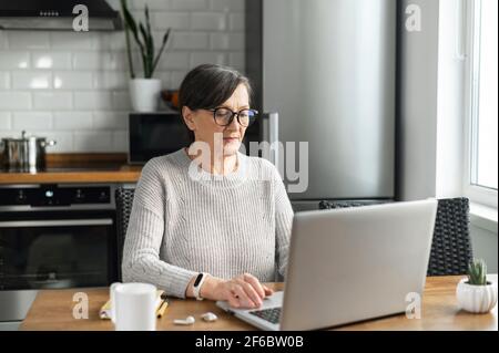 Retired woman using laptop for remote work, modern elderly female in eyewear sits at the kitchen desk and typing emails, conducts business correspondence on the distance Stock Photo