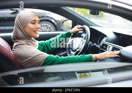 Cheerful muslim woman driving car, checking air conditioning Stock Photo