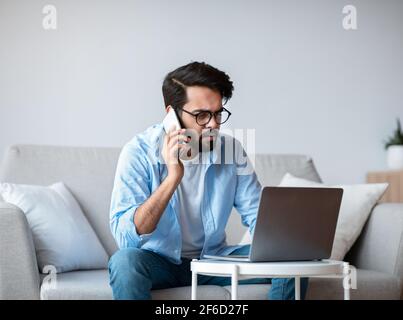 Busy Eastern Freelancer Guy Talking On Phone And Using Laptop At Home Stock Photo