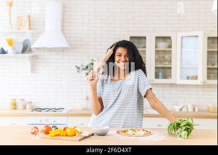 Young cheerful African American girl standing next to kitchen table and holding a rolling pin, concept of a woman in an everyday household routine, making cooking pizza, preparing dough and veggies Stock Photo