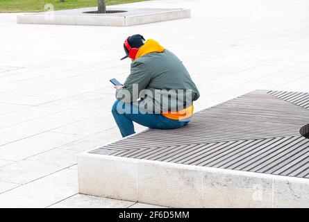 Mature man with headphones sitting on bench using phone. Full length portrait of caucasian modern young man. Stock Photo