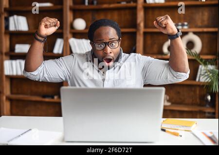 Overjoyed latin black businessman sitting at desk, looking at laptop screen, screaming, wow, celebrating success win, excited male guy feel euphoric receiving pleasant email message on computer. Stock Photo
