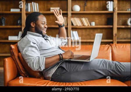Video call concept, pleasant senior latin unshaved man with dreadlocks, smiling, chatting greeting with family, using a laptop on the knees for video connection, say hi, glad to see, lying on couch Stock Photo
