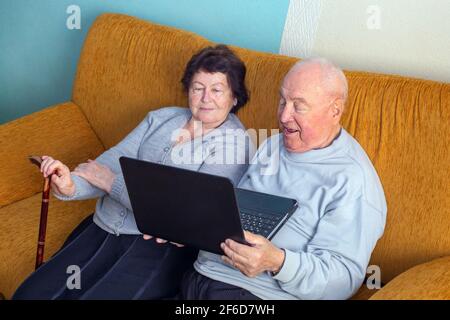Senior couple is talking online via video connection on the laptop on sofa in living room. Having nice time with family and friends via video call Stock Photo