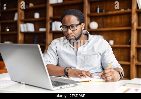Business planning, e-learning concept, casual african american business man writing on notebook and working using laptop computer in modern office, man studying online course or lecture via laptop, Stock Photo