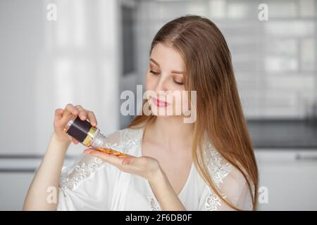 A beautiful woman in white clothes shows pouring out several tablets of vitamins dietary supplements from a jar in her kitchen. Stock Photo