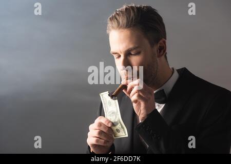 young businessman lighting cigar with hundred dollar banknote on grey background with smoke Stock Photo