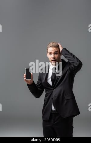 shocked businessman touching head while holding cellphone with blank screen isolated on grey Stock Photo