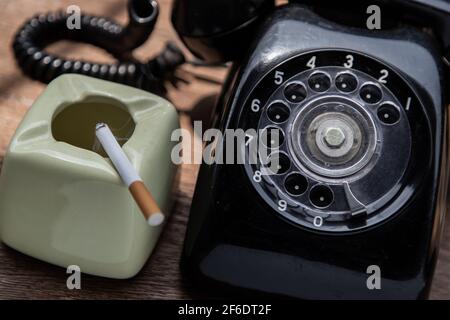 Cigarette that were burning placed on greyish green ashtray and Old black retro rotary telephone on old wooden rustic table. Copy space, Selective foc Stock Photo