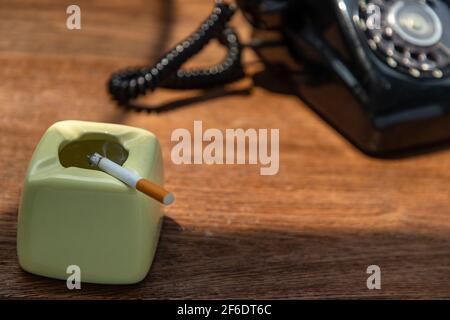 Cigarette that were burning placed on greyish green ashtray in front of Old black retro rotary telephone on old wooden rustic table. Copy space, Selec Stock Photo